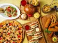 Obfity stół z jedzeniem fast food: pizza, burgery, frytki, kanapki, sałatka i panierowane przekąski.