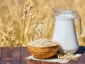 Mleko owsiane w dzbanku i miska płatków owsianych na tle łanu zbóż. Czy mleko owsiane ma gluten? Pytanie, które nurtuje wielu.