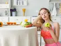 Młoda kobieta z miarką krawiecką na szyi trzyma jabłko, rozważając dietę 1100 kcal. Na stole warzywa i owoce.