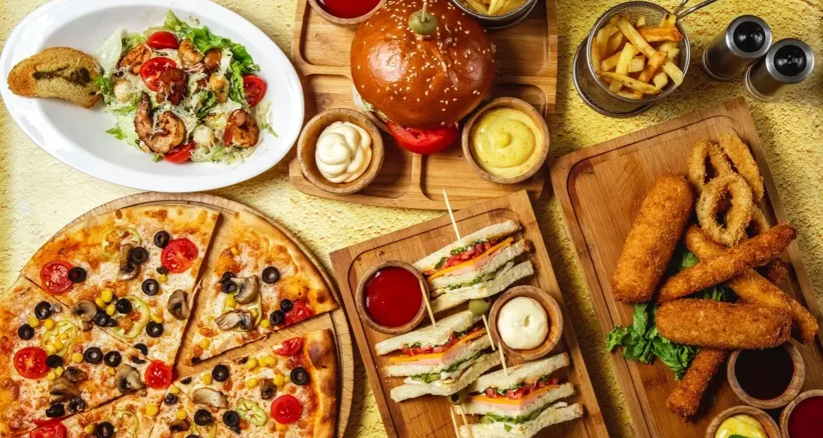 Obfity stół z jedzeniem fast food: pizza, burgery, frytki, kanapki, sałatka i panierowane przekąski.