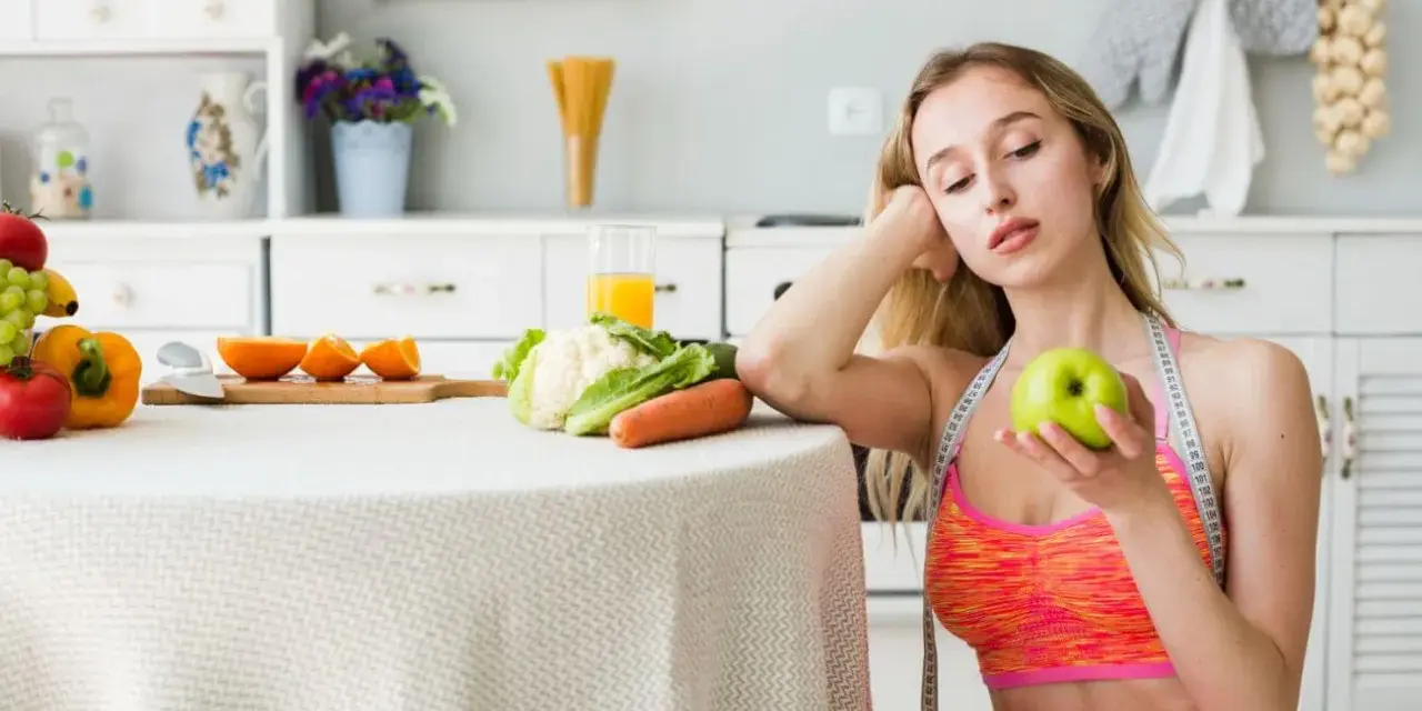 Młoda kobieta z miarką krawiecką na szyi trzyma jabłko, rozważając dietę 1100 kcal. Na stole warzywa i owoce.