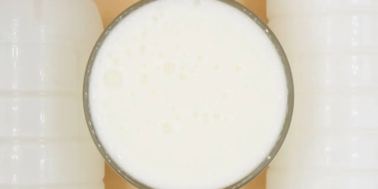 Szklanka pełna pianistego kefiru, otoczona białymi butelkami. Idealny moment, kiedy pić kefir, to teraz!