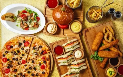 Obfity stół z jedzeniem fast food: pizza, burgery, frytki, kanapki, sałatka i panierowane przekąski.