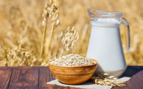 Mleko owsiane w dzbanku i miska płatków owsianych na tle łanu zbóż. Czy mleko owsiane ma gluten? Pytanie, które nurtuje wielu.