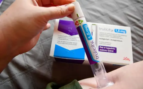 Ręka trzymająca pen do podawania zastrzyków na cukrzycę Trulicity 1,5 mg. W tle pudełko leku.