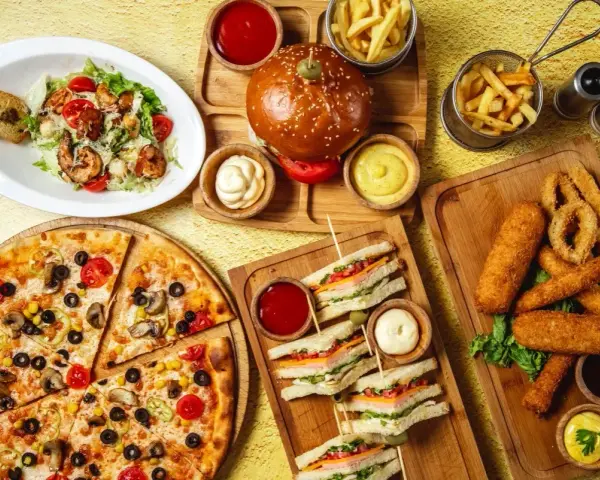 Obfity stół z jedzeniem fast food: pizza, burgery, frytki, kanapki, sałatka i panierowane przekąski.