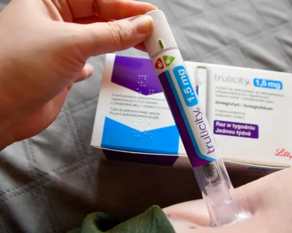 Ręka trzymająca pen do podawania zastrzyków na cukrzycę Trulicity 1,5 mg. W tle pudełko leku.