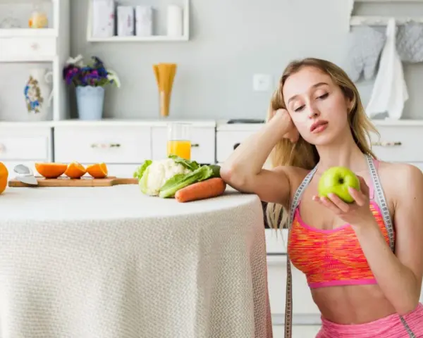 Młoda kobieta z miarką krawiecką na szyi trzyma jabłko, rozważając dietę 1100 kcal. Na stole warzywa i owoce.