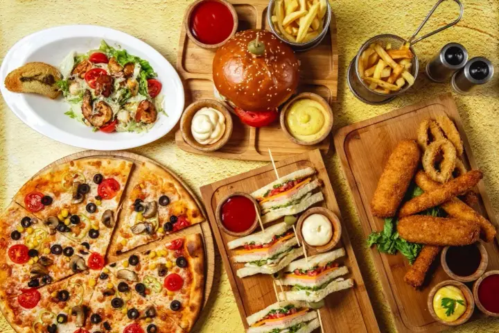 Obfity stół z jedzeniem fast food: pizza, burgery, frytki, kanapki, sałatka i panierowane przekąski.