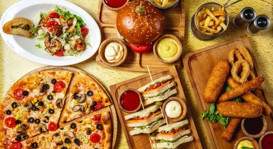 Obfity stół z jedzeniem fast food: pizza, burgery, frytki, kanapki, sałatka i panierowane przekąski.