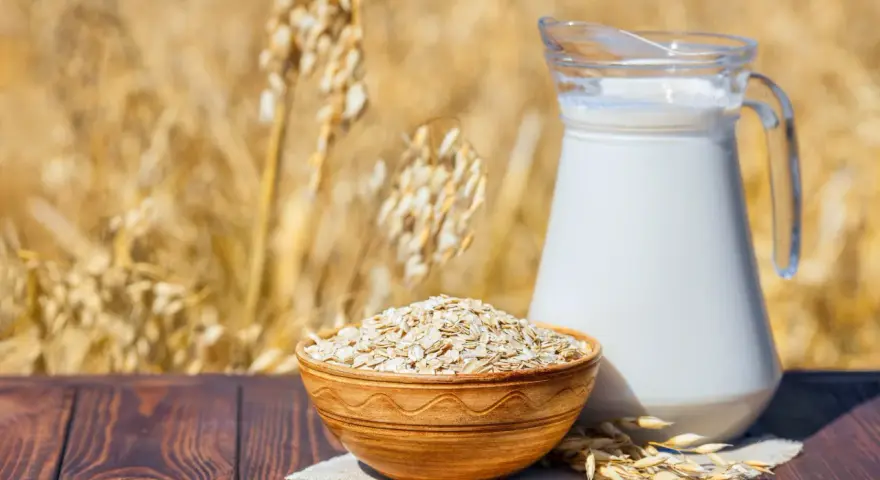 Mleko owsiane w dzbanku i miska płatków owsianych na tle łanu zbóż. Czy mleko owsiane ma gluten? Pytanie, które nurtuje wielu.