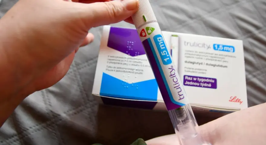 Ręka trzymająca pen do podawania zastrzyków na cukrzycę Trulicity 1,5 mg. W tle pudełko leku.