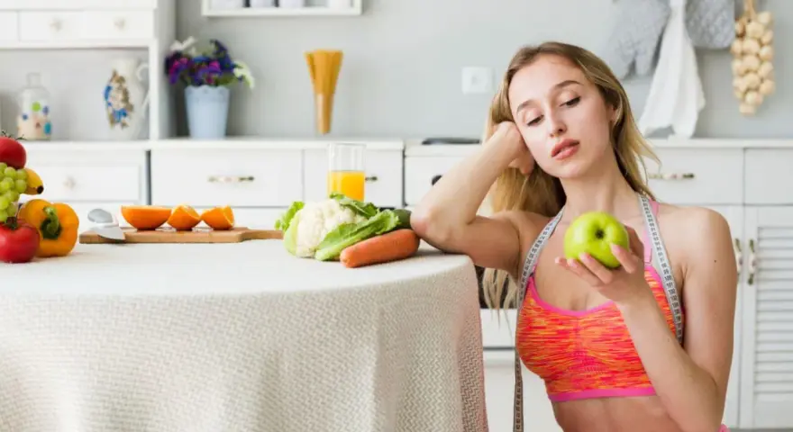 Młoda kobieta z miarką krawiecką na szyi trzyma jabłko, rozważając dietę 1100 kcal. Na stole warzywa i owoce.