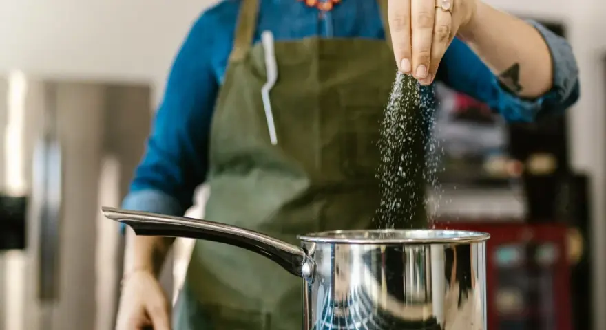 Kobieta w fartuchu dodaje sól do garnka, zastanawiając się, ile sodu dziennie jest bezpieczne.
