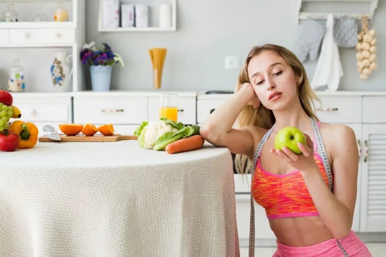 Kobieta z miarką krawiecką na szyi trzyma jabłko, obok stół z owocami i warzywami. Dieta 1100 kcal to wyzwanie.