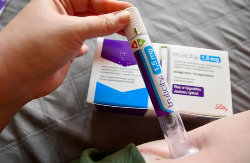Ręka trzymająca pen do podawania zastrzyków na cukrzycę Trulicity 1,5 mg. W tle pudełko leku.